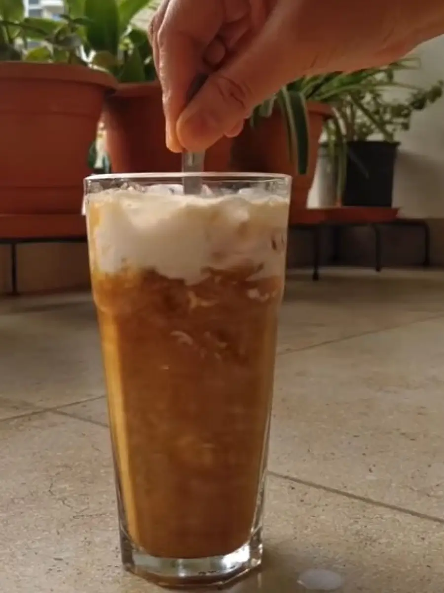 Hand stirring a tall glass of Sugar Free Coconut Milk Vietnamese Coffee with ice and creamy foam on top, indoor setting with potted plants in the background.